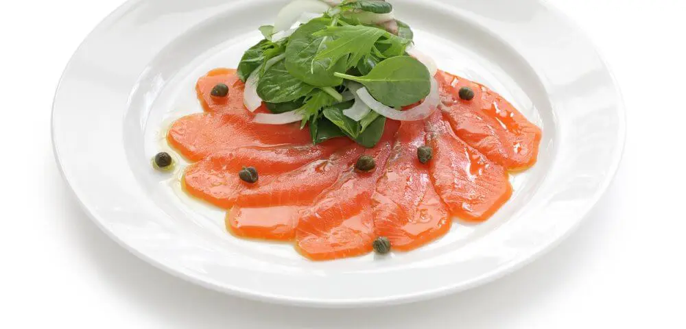 Carpaccio de saumon, présenté sous forme de coulis dans une assiette blanche, avec quelques feuilles de salade dessus.